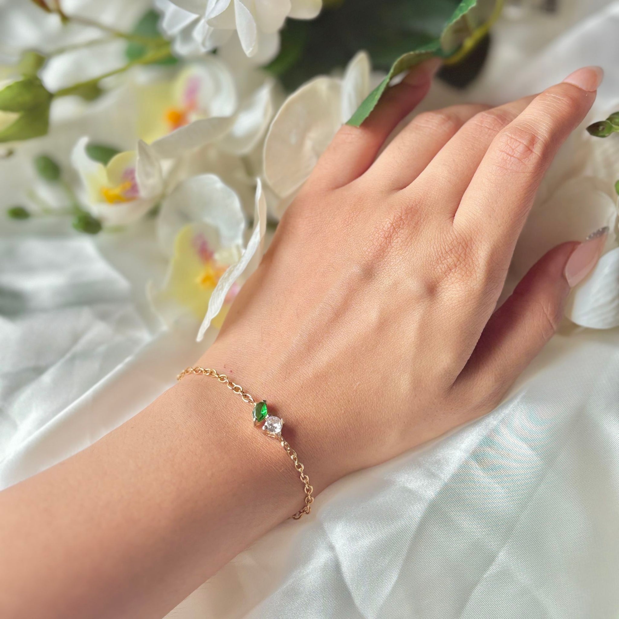 Hand wearing a gold bracelet with green and pink stones against a floral background